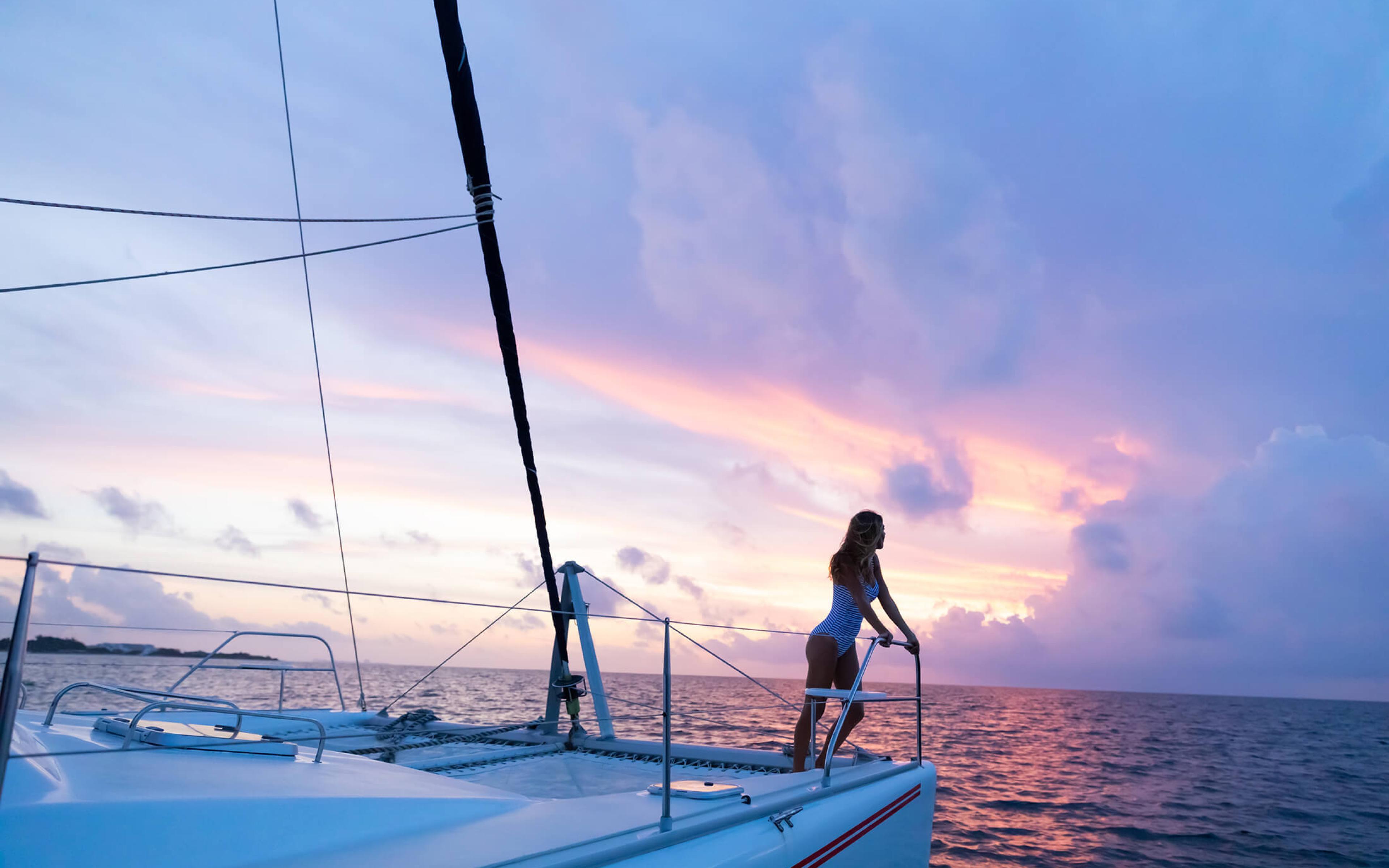 A woman leans on the bow of a catamaran at sunset